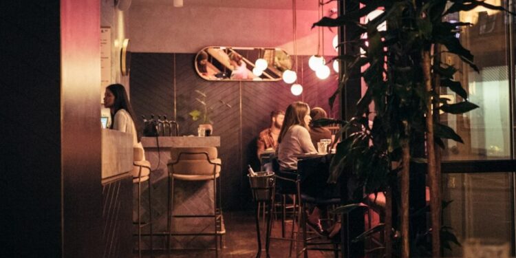 Members clubs London worth joining - GUSTOwebzine Café interior at night with warm pink lighting, patrons seated at tables near a bar; leafy plants in the foreground.