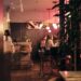 Café interior at night with warm pink lighting, patrons seated at tables near a bar; leafy plants in the foreground.