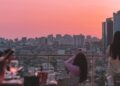 People on a rooftop terrace at sunset, overlooking a dense city skyline with a pink-orange sky.