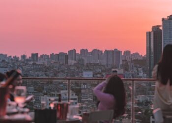 People on a rooftop terrace at sunset, overlooking a dense city skyline with a pink-orange sky.