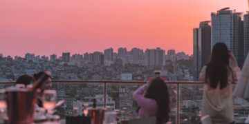 People on a rooftop terrace at sunset, overlooking a dense city skyline with a pink-orange sky.