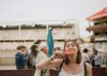 Young woman in a white top bites into a chicken wing at an outdoor fair, with a man in a feather headdress and other attendees in the background.