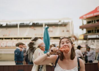 Young woman in a white top bites into a chicken wing at an outdoor fair, with a man in a feather headdress and other attendees in the background.