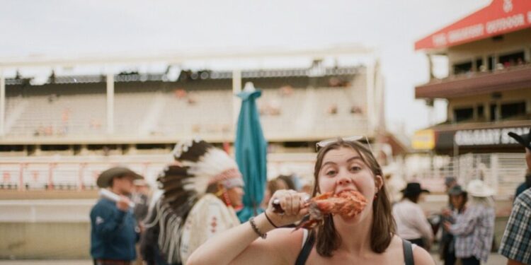 Young woman in a white top bites into a chicken wing at an outdoor fair, with a man in a feather headdress and other attendees in the background.