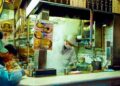 Soba frío y tofu helado en Tokio - GUSTOwebzine Mostrador abierto de tienda de fideos japoneses con un chef de sombrero blanco y mascarilla cocinando entre vapores ascendentes mientras los clientes comen en la barra a la izquierda.