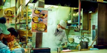 Soba frío y tofu helado en Tokio - GUSTOwebzine Mostrador abierto de tienda de fideos japoneses con un chef de sombrero blanco y mascarilla cocinando entre vapores ascendentes mientras los clientes comen en la barra a la izquierda.