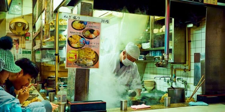 Soba frío y tofu helado en Tokio - GUSTOwebzine Mostrador abierto de tienda de fideos japoneses con un chef de sombrero blanco y mascarilla cocinando entre vapores ascendentes mientras los clientes comen en la barra a la izquierda.