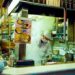 Mostrador abierto de tienda de fideos japoneses con un chef de sombrero blanco y mascarilla cocinando entre vapores ascendentes mientras los clientes comen en la barra a la izquierda.