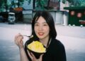 Young woman outdoors, smiling while eating a bowl of mango shaved ice with a spoon.