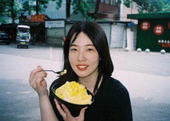 Young woman outdoors, smiling while eating a bowl of mango shaved ice with a spoon.