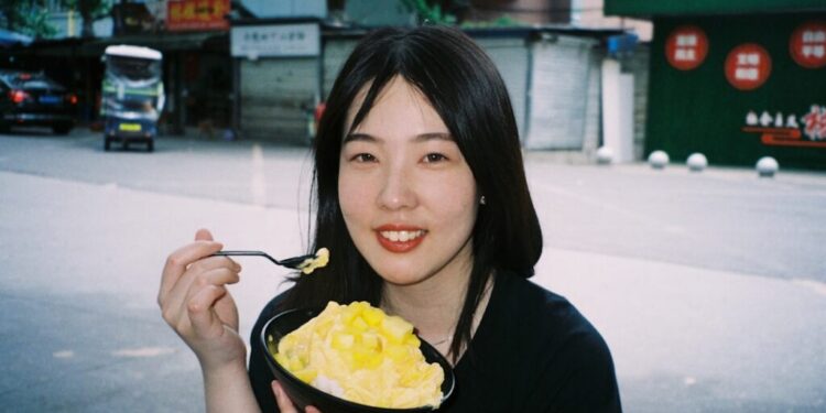 Young woman outdoors, smiling while eating a bowl of mango shaved ice with a spoon.