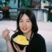 Young woman outdoors, smiling while eating a bowl of mango shaved ice with a spoon.