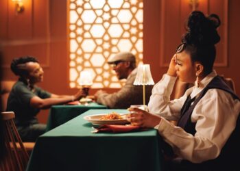 Woman with an elaborate bun sits at a green-clothed table in a warmly lit restaurant, resting her head on her hand as she looks away; plate of pasta in front.