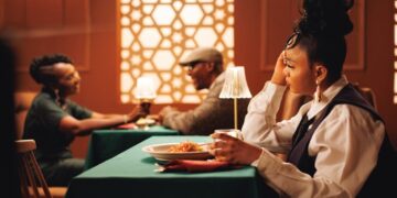 Mujer con un peinado elaborado sentada en una mesa cubierta con mantel verde en un restaurante cálidamente iluminado, apoyando su cabeza en su mano mientras mira hacia otro lado; plato de pasta frente a ella.