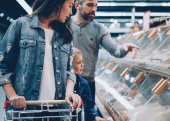 Mother pushes a shopping cart with her young son nearby as the father points to pastries in a glass bakery display case in a store.