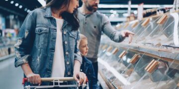 Madre che spinge un carrello della spesa con suo figlio piccolo nelle vicinanze mentre il padre indica i dolci in una vetrina di una panetteria in un negozio.