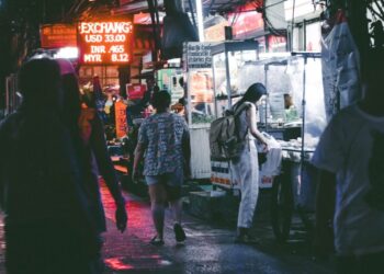 Night market street at night with neon signs; a woman with a backpack buys food from a stall as other pedestrians walk by, wet pavement reflecting lights.