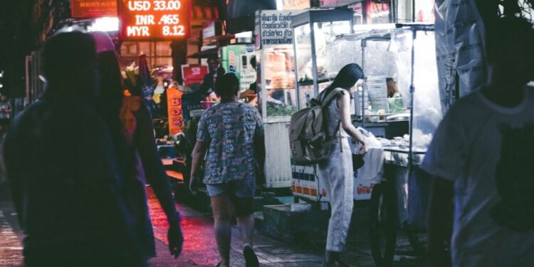 Glutenfreier Hawker-Center-Guide Singapur - GUSTOwebzine Nachtmarktstraße nachts mit Neonlichtern; eine Frau mit Rucksack kauft Essen an einem Stand, während andere Fußgänger vorbeigehen, nasse Gehweg spiegelt Lichter wider.