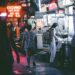 Night market street at night with neon signs; a woman with a backpack buys food from a stall as other pedestrians walk by, wet pavement reflecting lights.
