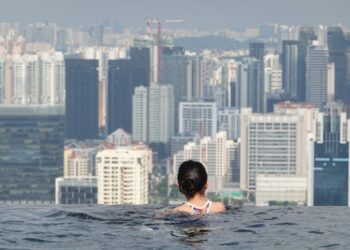 Back view of a person with dark hair in an infinity pool, facing a dense city skyline.