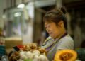 Depachika gluten-free meals in Tokyo - GUSTOwebzine Market vendor, a woman in a striped shirt looking at her phone while standing near piles of produce including garlic and ginger in a busy market stall.