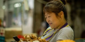 Depachika gluten-free meals in Tokyo - GUSTOwebzine Market vendor, a woman in a striped shirt looking at her phone while standing near piles of produce including garlic and ginger in a busy market stall.