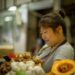 Market vendor, a woman in a striped shirt looking at her phone while standing near piles of produce including garlic and ginger in a busy market stall.