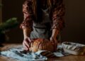 Wheat-free bread traditions across the EU - GUSTOwebzine Person in a floral blouse shapes a round rustic loaf of bread on a striped towel on a wooden table