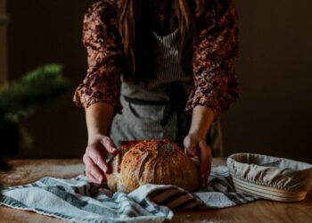 Persoon in een bloemetjesblouse vormt een rond rustiek brood op een gestreepte theedoek op een houten tafel