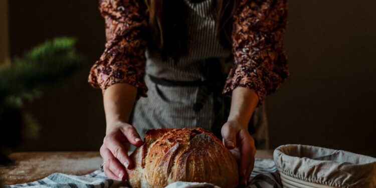 Wheat-free bread traditions across the EU - GUSTOwebzine Person in a floral blouse shapes a round rustic loaf of bread on a striped towel on a wooden table