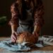 Person in a floral blouse shapes a round rustic loaf of bread on a striped towel on a wooden table
