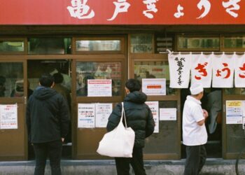 Mensen staan in de rij voor een Japans eethuis met witte gordijnen en rode uithangborden boven de deur, en posters op de ramen.