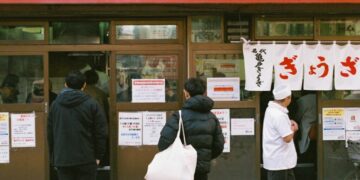 Singapore dining resets before September rush - GUSTOwebzine People stand in line outside a Japanese eatery with white curtain banners and red signage above the door, posters on the windows.