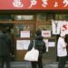 People stand in line outside a Japanese eatery with white curtain banners and red signage above the door, posters on the windows.