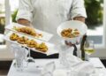 Autumn menus in Dubai worth booking now - GUSTOwebzine Chef in a white uniform presents two rectangular plates of bite-sized appetizers and a white bowl of fried chicken, on a white-tablecloth setup with a wine glass and bottle in the background.