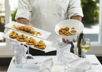 Een chef-kok in een wit uniform presenteert twee rechthoekige borden met hapjes en een witte kom met gefrituurde kip, op een tafel met een wit kleed, een wijnglas en een fles op de achtergrond.