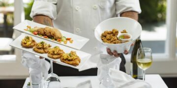 Autumn menus in Dubai worth booking now - GUSTOwebzine Chef in a white uniform presents two rectangular plates of bite-sized appetizers and a white bowl of fried chicken, on a white-tablecloth setup with a wine glass and bottle in the background.