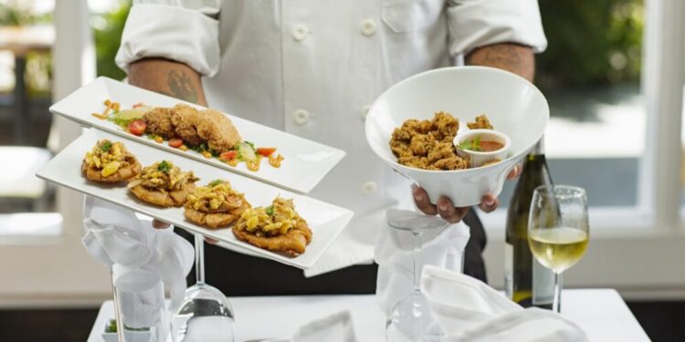 Autumn menus in Dubai worth booking now - GUSTOwebzine Chef in a white uniform presents two rectangular plates of bite-sized appetizers and a white bowl of fried chicken, on a white-tablecloth setup with a wine glass and bottle in the background.