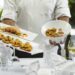 Autumn menus in Dubai worth booking now - GUSTOwebzine Chef in a white uniform presents two rectangular plates of bite-sized appetizers and a white bowl of fried chicken, on a white-tablecloth setup with a wine glass and bottle in the background.