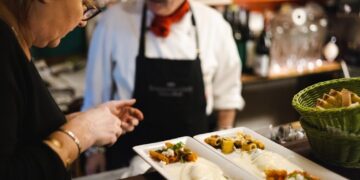 Two staff members in a kitchen plate upscale dishes at a counter, with a green bread basket nearby and plated courses in the foreground