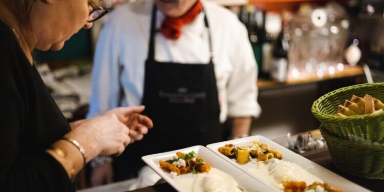 Two staff members in a kitchen plate upscale dishes at a counter, with a green bread basket nearby and plated courses in the foreground