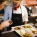 Two staff members in a kitchen plate upscale dishes at a counter, with a green bread basket nearby and plated courses in the foreground