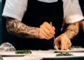 A professional chef adjusting salt acid and seasoning in a large pot in a busy restaurant kitchen
