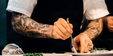 A professional chef adjusting salt acid and seasoning in a large pot in a busy restaurant kitchen