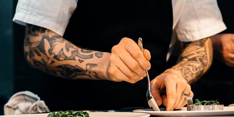 A professional chef adjusting salt acid and seasoning in a large pot in a busy restaurant kitchen