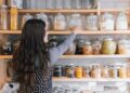 Woman with long wavy hair reaching for a glass jar on a wooden pantry shelf filled with jars of grains and spices.