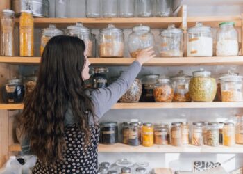 Spice pantry built on Indias 6 regions - GUSTOwebzine Woman with long wavy hair reaching for a glass jar on a wooden pantry shelf filled with jars of grains and spices.