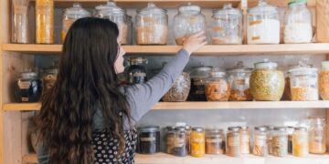 Woman with long wavy hair reaching for a glass jar on a wooden pantry shelf filled with jars of grains and spices.