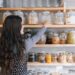 Spice pantry built on Indias 6 regions - GUSTOwebzine Woman with long wavy hair reaching for a glass jar on a wooden pantry shelf filled with jars of grains and spices.
