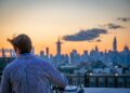 DJ on a rooftop deck mixing music as the city skyline glows at sunset behind him, wearing a striped shirt.